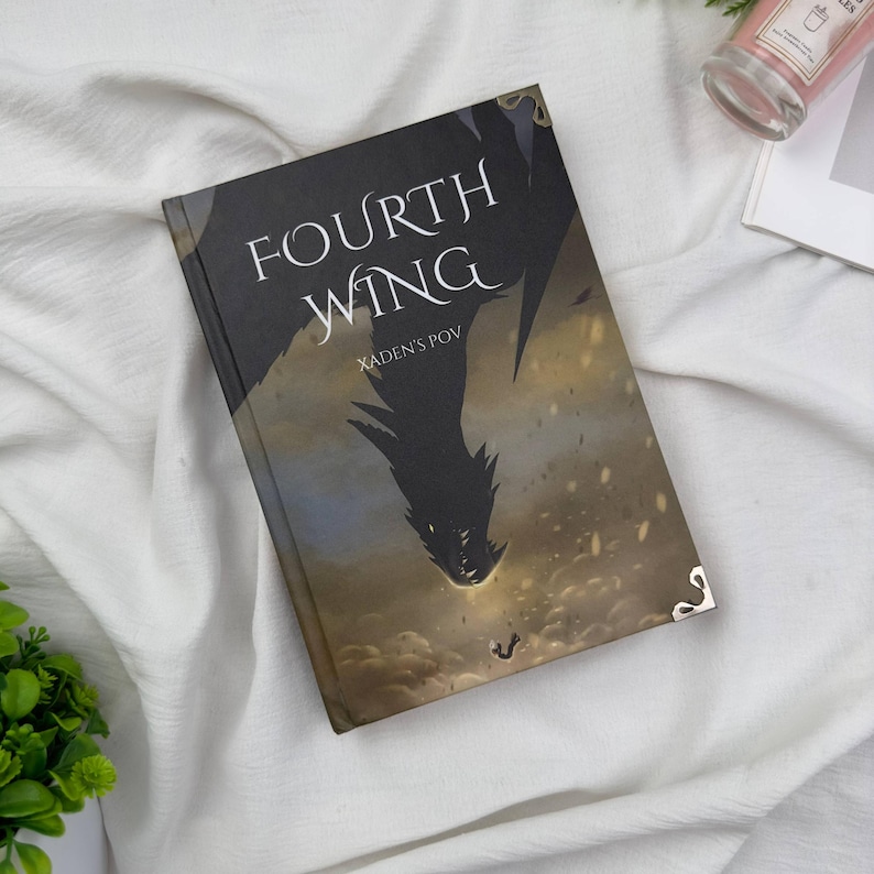 May include: A hardcover book titled "FOURTH WING" with "XADEN'S POV" below the title. The cover features a dark silhouette of a dragon against a cloudy, golden background. The book has gold corner protectors.