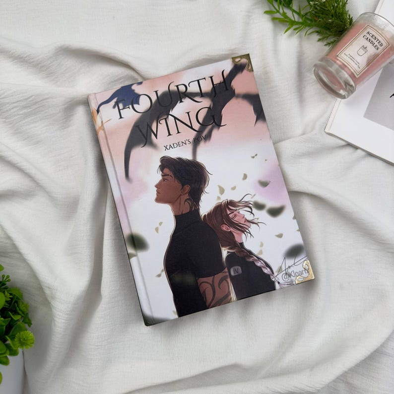 May include: A hardcover book titled "Fourth Wing" with a fantasy illustration of two people. The cover art features a man and woman with a dragon silhouette and floral accents. The book is on a white surface.