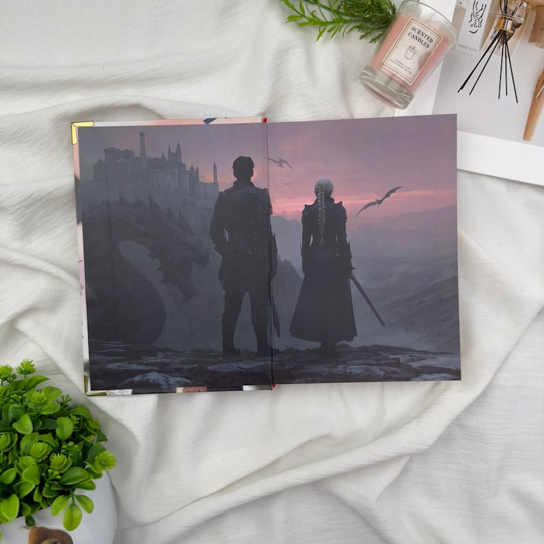 May include: Open journal with a fantasy illustration. The artwork depicts two figures standing on a cliff, overlooking a castle and a dragon, with a sunset sky. The journal has gold corner protectors.