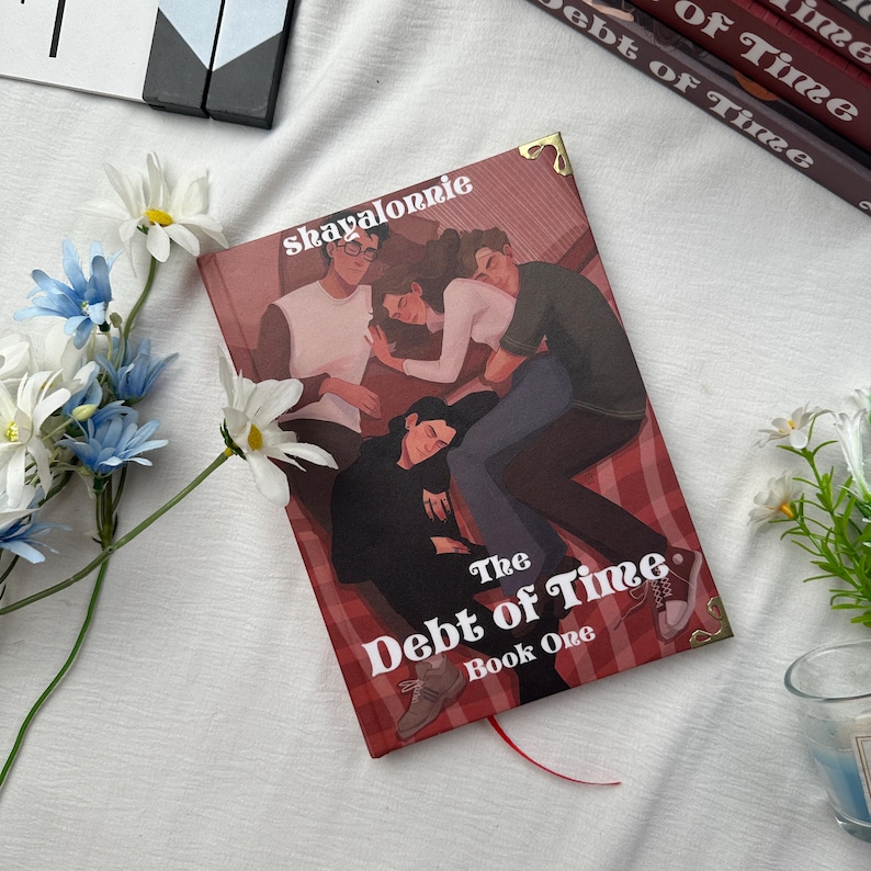 May include: A hardcover book titled "The Debt of Time Book One" with a cover illustration of people. The book has a red bookmark and gold corner accents. The author's name, "shayalorrie", is printed at the top. The book is surrounded by flowers.