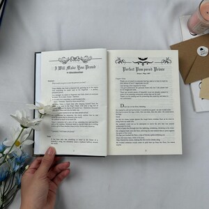 May include: An open book with black covers, displaying text on cream-colored pages. The left page reads "I Will Make You Proud" with decorative flourishes. The right page reads "Perfect Pampered Prince". A hand holds the book, with flowers and a candle in the background.