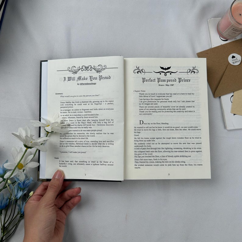May include: An open book with black covers, displaying text on cream-colored pages. The left page reads "I Will Make You Proud" with decorative flourishes. The right page reads "Perfect Pampered Prince". A hand holds the book, with flowers and a candle in the background.