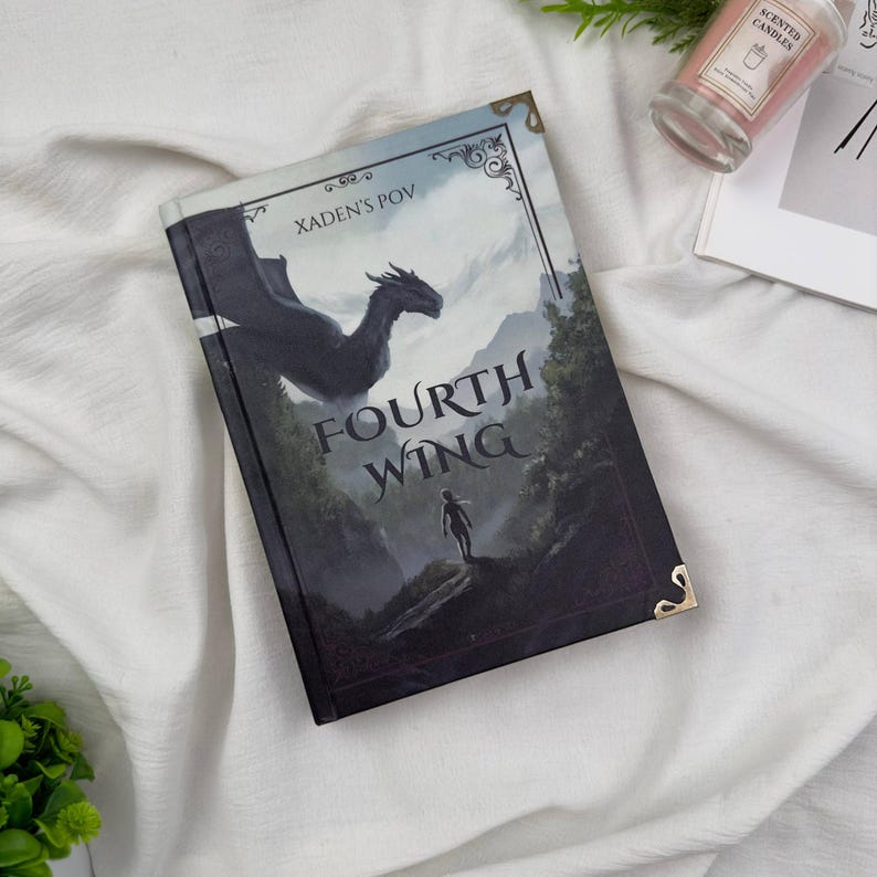 May include: A hardcover book titled "Fourth Wing" with a dragon illustration and the text "Xaden's POV." The cover features a dragon, a figure, and a mountain landscape. The book has gold corner protectors.