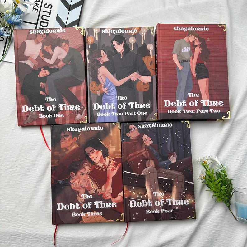 May include: A collection of five books titled "The Debt of Time" with various cover illustrations. The covers feature couples in romantic settings, with text including "shayalonnie" and book numbers. The books are arranged on a white surface.
