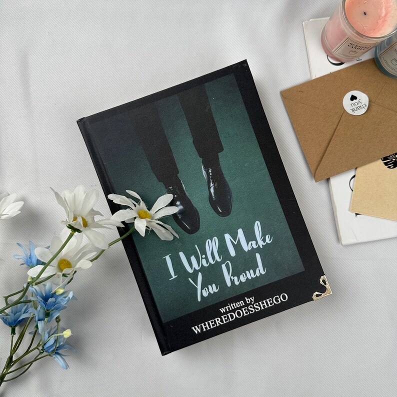 May include: A hardcover book titled "I Will Make You Proud" with a dark green cover featuring an image of legs and dress shoes. The author's name, WHEREDOESSHEGO, is also visible. The book is decorated with white and blue flowers.