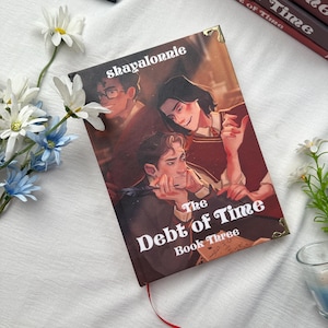 May include: A hardcover book titled "The Debt of Time, Book Three" with a cover illustration of three figures. The author's name, "shayalornie," is printed at the top. The book has a red ribbon bookmark and gold corner accents.