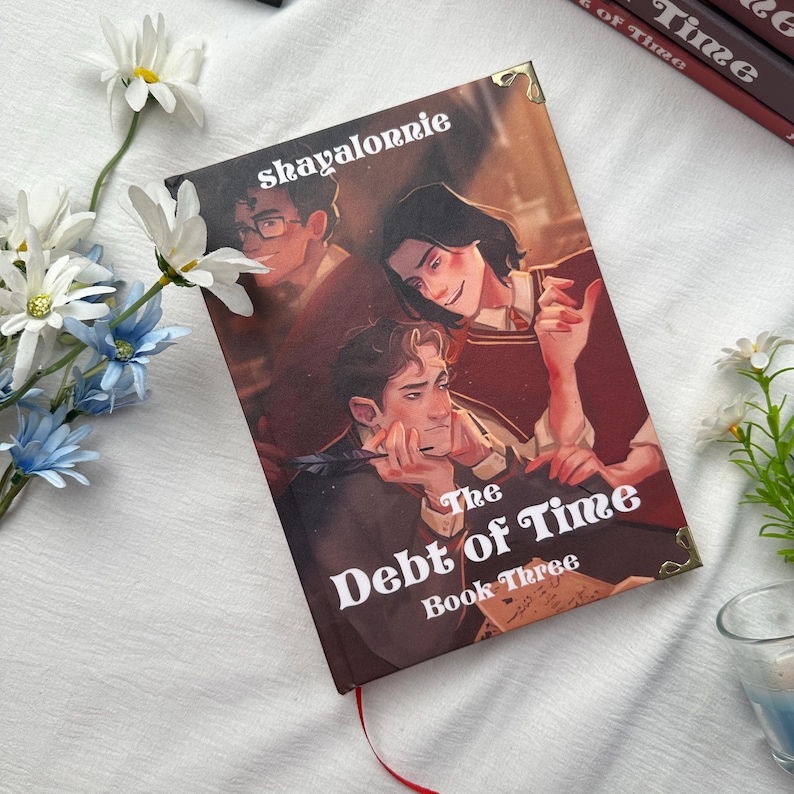 May include: A hardcover book titled "The Debt of Time, Book Three" with a cover illustration of three figures. The author's name, "shayalornie," is printed at the top. The book has a red ribbon bookmark and gold corner accents.