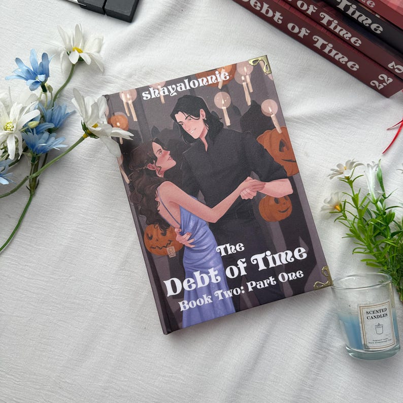 May include: A book titled "The Debt of Time: Book Two: Part One" features a couple dancing on the cover. The cover art includes a woman in a blue dress and a man in black, with pumpkin decorations. The author's name, shayalonnie, is also visible.