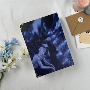 May include: A hardcover book with a dark blue cover featuring an illustration of a man and woman. The cover art has a dramatic, romantic theme. Gold corner protectors add a decorative touch. The book is on a white surface with flowers and other items.