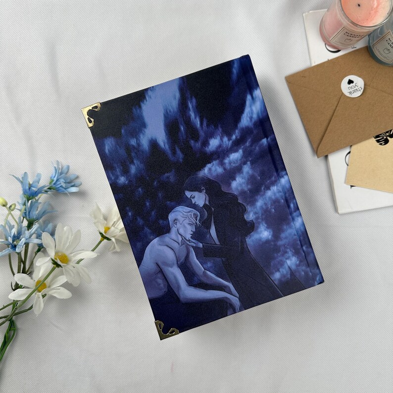May include: A hardcover book with a dark blue cover featuring an illustration of a man and woman. The cover art has a dramatic, romantic theme. Gold corner protectors add a decorative touch. The book is on a white surface with flowers and other items.