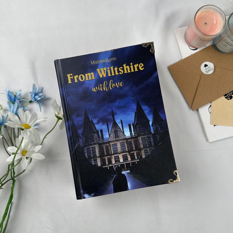 May include: A hardcover book titled "From Wiltshire with love" by MistressLynn. The cover features a dark blue night sky with a castle illustration. Gold corner protectors and text accents the cover. The book is placed on a white surface with flowers and candles.