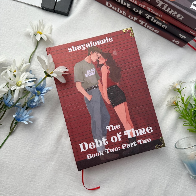 May include: A hardcover book titled "The Debt of Time: Book Two: Part Two" with a red brick wall design. The cover features an illustration of a couple. The author's name, "shayalonnie", is printed at the top.