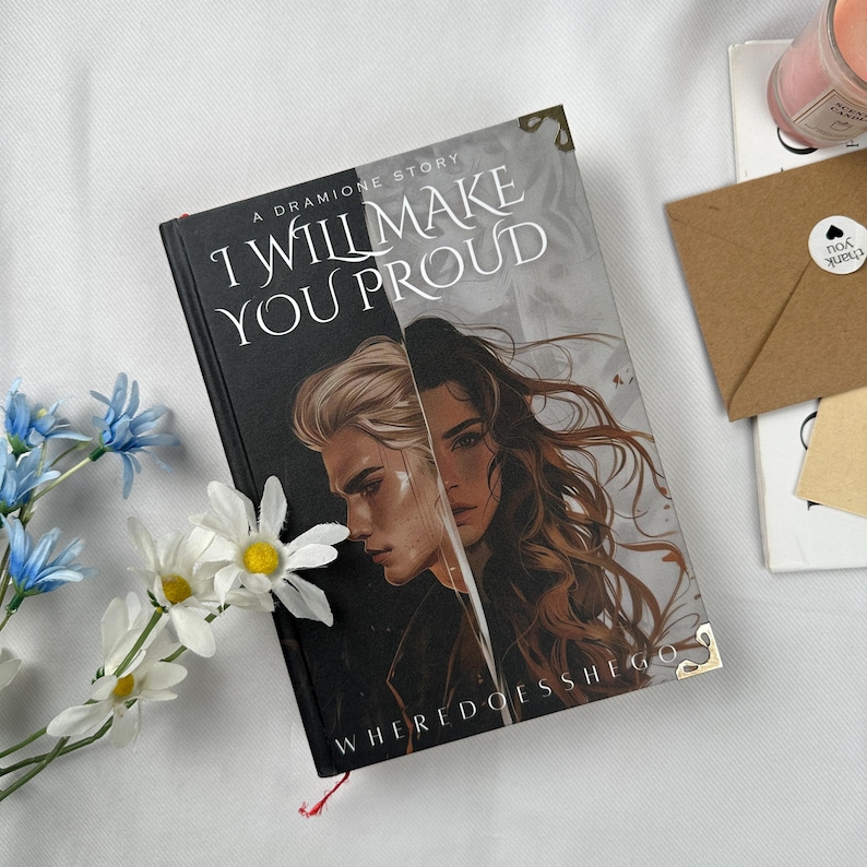 May include: A book titled "I Will Make You Proud" with a cover featuring two illustrated faces. The book has a black spine and the text "WHERE DOES SHE GO" at the bottom. A sprig of white and blue flowers is next to the book.