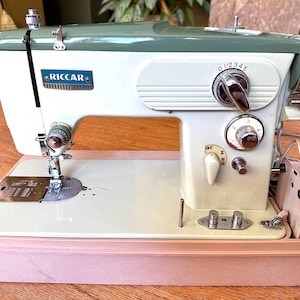 May include: Vintage Riccar sewing machine in a light green and white color scheme. The machine features a silver needle, dials, and a pink base. The Riccar logo is visible on the front. The machine is in a light pink case.