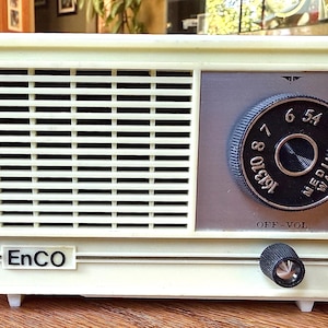 May include: A white vintage EnCO radio with a black dial and knobs. The dial has numbers 5.4, 6, 7, 8, 9, 10, and 12. The radio has a black speaker grille and the words "OFF-VOL" and "MEDIUM WAVE" are printed on the dial.