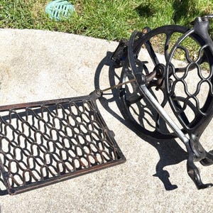 May include: An antique black cast iron sewing machine base with a decorative openwork design. The base includes a large flywheel, a connecting rod, and a rectangular, patterned plate. The machine is set on a concrete surface.