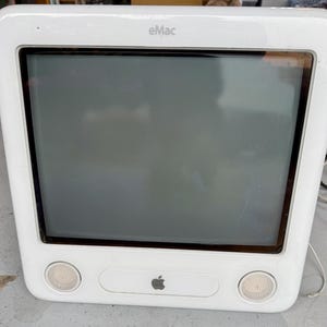 May include: White eMac computer with a dark screen, two speaker grills, and the Apple logo. The computer has the word "eMac" printed on the top. The computer is a vintage model.