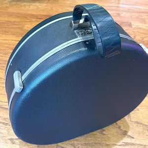 May include: A round, black, hardshell carrying case with a textured exterior, a silver-toned metal trim, and a dark handle featuring a decorative embossed pattern. The case includes silver-toned metal latches and hinges along the side and top.