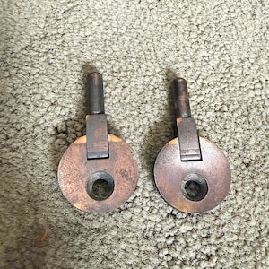 May include: Two matching metal furniture pulls with a weathered copper finish. Each pull features a circular plate with a central screw hole and a vertical post extending from the top, designed for attachment to a drawer or cabinet front.