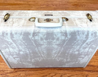 May include: A vintage white hard-sided suitcase with a marbled pattern and gold accents. The suitcase has a white handle and two gold latches.
