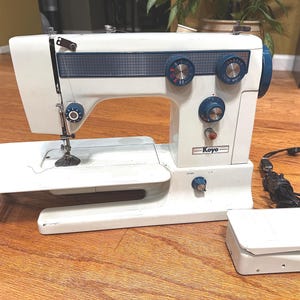 Pode incluir: Máquina de costura Koyo branca vintage com detalhes azuis. A máquina tem vários mostradores e uma agulha, com uma mesa de extensão branca e um pedal. Sobre uma superfície de madeira.