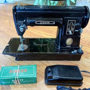 Könnte beinhalten: Eine schwarze Singer-Nähmaschine mit Goldverzierungen, einem Stichlängenregler und einem runden Emblem. Dazu gehören eine grüne rechteckige Schachtel mit der Aufschrift Singer Sewing Machine Attachments und ein schwarzes Fußpedal.