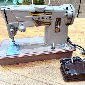 May include: A vintage Singer sewing machine in a neutral color palette. The machine features the Singer logo in gold and brown, with a foot pedal and power cord. The sewing machine is set on a brown base with a light blue area.