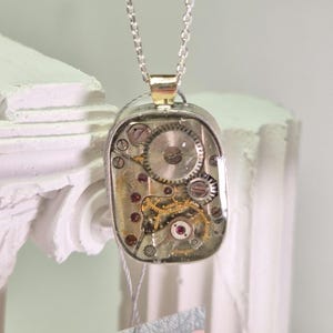 May include: A silver pendant necklace with a rectangular, clear casing displaying intricate watch gears and ruby-coloured jewels. The pendant hangs from a delicate chain, creating a unique, industrial-inspired accessory.