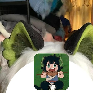 May include: A pair of faux fur animal ears featuring black outer edges and lime green inner sections with a scalloped design. The ears are attached to a white faux fur base. A square graphic shows a cartoon character eating a fish.