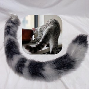 Puede incluir: Una cola de gato de piel sintética en tonos grises y blancos, curvada en forma de media luna. La cola tiene una textura esponjosa y una punta gris oscuro. Un gato es visible en el fondo.