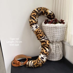 May include: A brown and black faux fur tiger tail with a white stripe down the middle. The tail is 70 cm long and comes with a brown leather belt. The tail is curled up and resting on a white fabric surface.