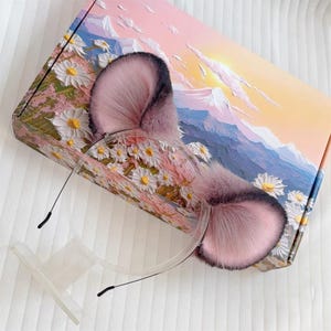 May include: A pair of pink and gray faux fur cat ears on a clear headband. The ears are fluffy and detailed. The headband is resting on a white stand, with a box in the background featuring a floral and mountain landscape.