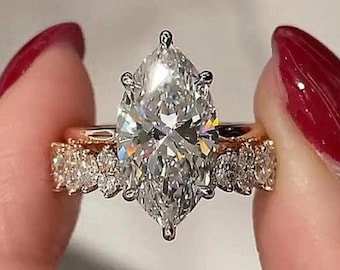 May include: A marquise-cut diamond engagement ring with a rose gold band. The ring features a large, clear, marquise-cut diamond as the centerpiece, with smaller diamonds set along the band. The ring is held between two fingers with red nail polish.