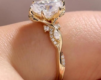Puede incluir: Un anillo de compromiso de oro con un gran diamante de talla ovalada. El anillo tiene un diseño floral con delicados detalles de hojas y vides. Pequeñas piedras de acento están engastadas a lo largo de la banda.