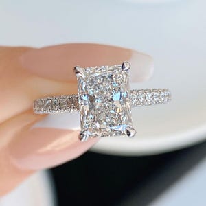 May include: A rectangular-cut diamond engagement ring. The large central diamond is set in a platinum or white gold band. The band features smaller, pavé-set diamonds, enhancing the sparkle. The ring is held against a soft, blurred background.