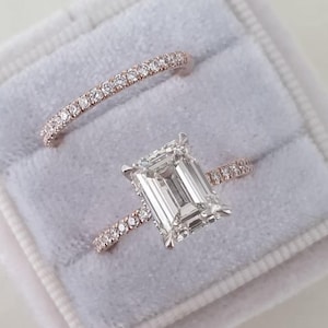 Puede incluir: Dos anillos de oro rosa en una caja de terciopelo gris. Uno tiene una gema clara rectangular grande. El otro es una banda delgada con pequeñas gemas claras. Para compromiso o boda.