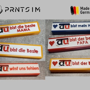 Peut inclure: Cinq plaques différentes imprimées en 3D avec un cœur rouge, le mot "du" en rouge et blanc, et un message en noir. Les plaques sont jaunes, rouges, bleues et blanches. Les messages sont "du bist die beste MAMA", "du bist mein Held", "du bist die Beste", "du bist der beste PAPA", et "du wirst uns fehlen".
