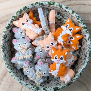 Op de afbeelding: Een kom gevuld met kleurrijke siliconen bedels in de vorm van katten. De bedels zijn in tinten oranje, grijs en roze.