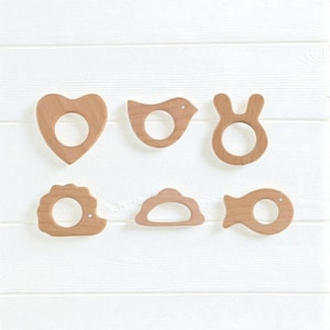 Peut inclure: Six hochets en bois pour bébés de différentes formes : un cœur, un oiseau, un lapin, un hérisson, un nuage et un poisson. Chaque hochet a une ouverture circulaire au centre. Les hochets sont en bois clair et sont disposés sur une surface en bois blanc.