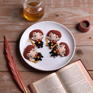 Könnte beinhalten: Vier Harry Potter-Charakterdekorationen auf einem weißen Teller. Jede Figur hat braunes Haar, eine Brille und einen Schal. Ein Glas mit gelber Flüssigkeit, eine kleine braune Schüssel, eine Feder und ein offenes Buch sind ebenfalls im Bild.