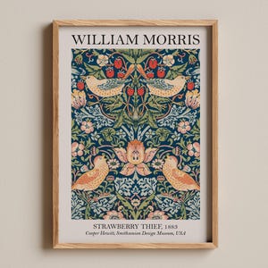 Op de afbeelding: Een ingelijste print van William Morris' "Strawberry Thief" ontwerp. Het kunstwerk toont een herhalend patroon van vogels, aardbeien en bladeren in blauwe, groene en perziktinten. De tekst "WILLIAM MORRIS" staat bovenaan en "STRAWBERRY THIEF, 1883" onderaan.