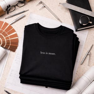 May include: A pile of black t-shirts, each bearing the white text "less is more". The shirts rest on a blueprint, surrounded by drafting tools and a colour swatch fan.