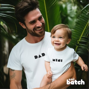 May include: A father and son wearing matching white t-shirts with black geometric text. The father's shirt says "DAD" and the son's shirt says "MINI".