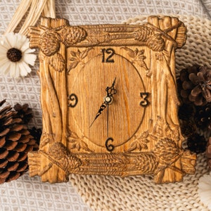 Könnte beinhalten: Eine quadratische Holzuhr mit hellbraunem Finish. Das Zifferblatt ist rund mit schwarzen Ziffern und Zeigern. Der Rahmen ist mit einem rustikalen Design aus Ästen, Tannenzapfen und Blättern geschnitzt. Die Uhr zeigt ungefähr 7:00 Uhr an.