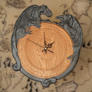 May include: A round clock with a bronze-colored face and black hands, encircled by two gray dragon figures. The clock face has an intricate design, and the dragons appear to be holding the clock. The background has a map design.