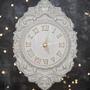 Puede incluir: Un reloj de pared blanco y ornamentado con números romanos y manecillas doradas. El reloj tiene un marco decorativo y con volutas. El fondo es oscuro con luces suaves y desenfocadas, creando una estética vintage.
