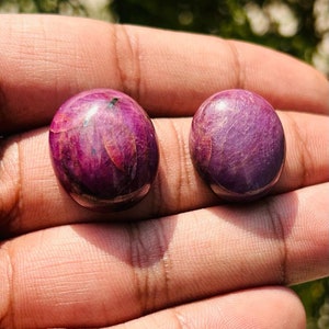 May include: Two oval-shaped, cabochon cut gemstones. The gemstones are a deep, rich purple color with a slight reddish hue. The stones are smooth and polished.