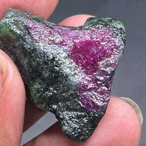 Puede incluir: Un espécimen de rubí y zoisita en bruto. La piedra presenta una sección de rubí magenta intenso contrastada con una matriz de zoisita verde oscuro y gris. La textura en bruto y sin pulir es visible.
