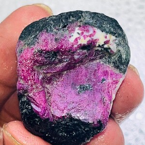Puede incluir: Una gema en bruto con un llamativo contraste de colores. La piedra presenta un núcleo magenta intenso, enmarcado por una capa exterior negra. Parches blancos y rosas claros añaden complejidad visual al espécimen mineral.