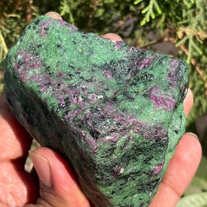 May include: A natural ruby zoisite stone, roughly cut. The stone has a vibrant green base colour with dark black and deep pink inclusions. The stone is held in a hand, with a blurred green background.